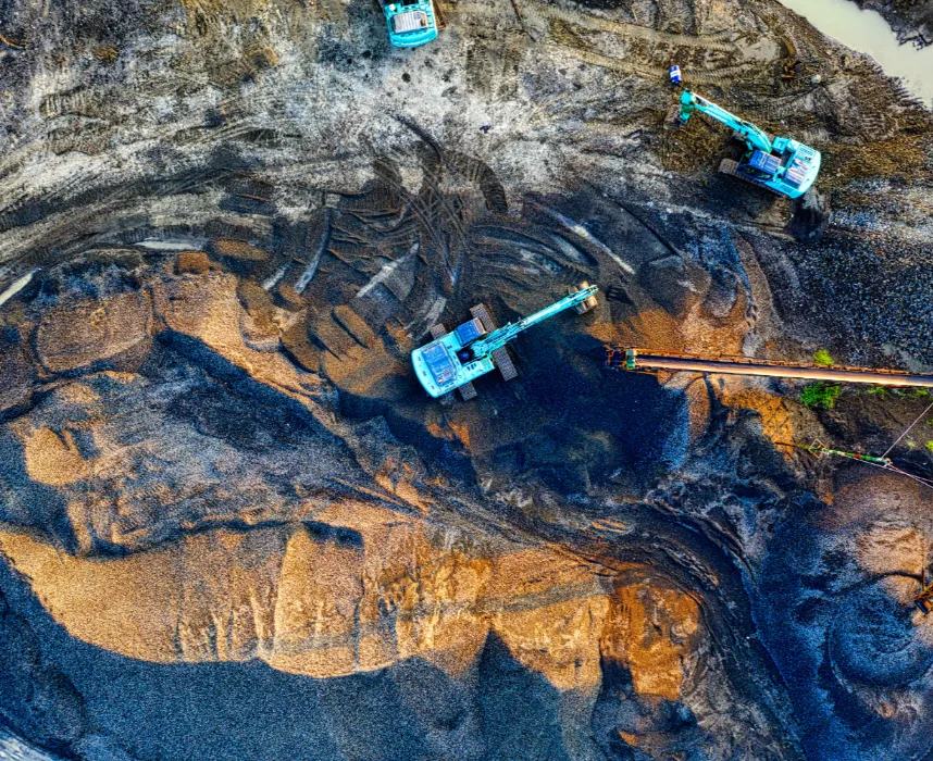 Aerial view of an open-pit mine with several teal-colored excavators and mining equipment working on piles of dark earth and golden-brown aggregate, showing tire tracks and a conveyor belt.